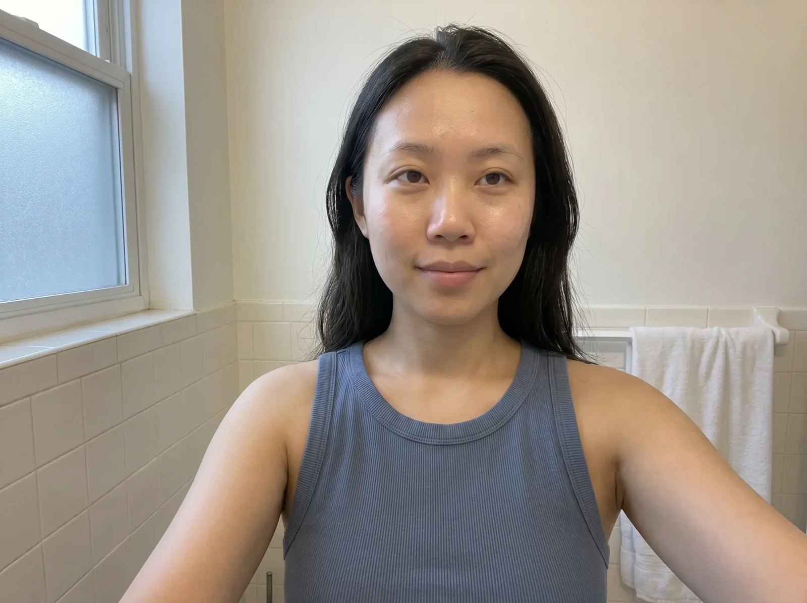 Selfie of the same young woman showing visibly reduced puffiness and brighter, more even skin, no makeup, wet hair, blue tank top, bathroom setting