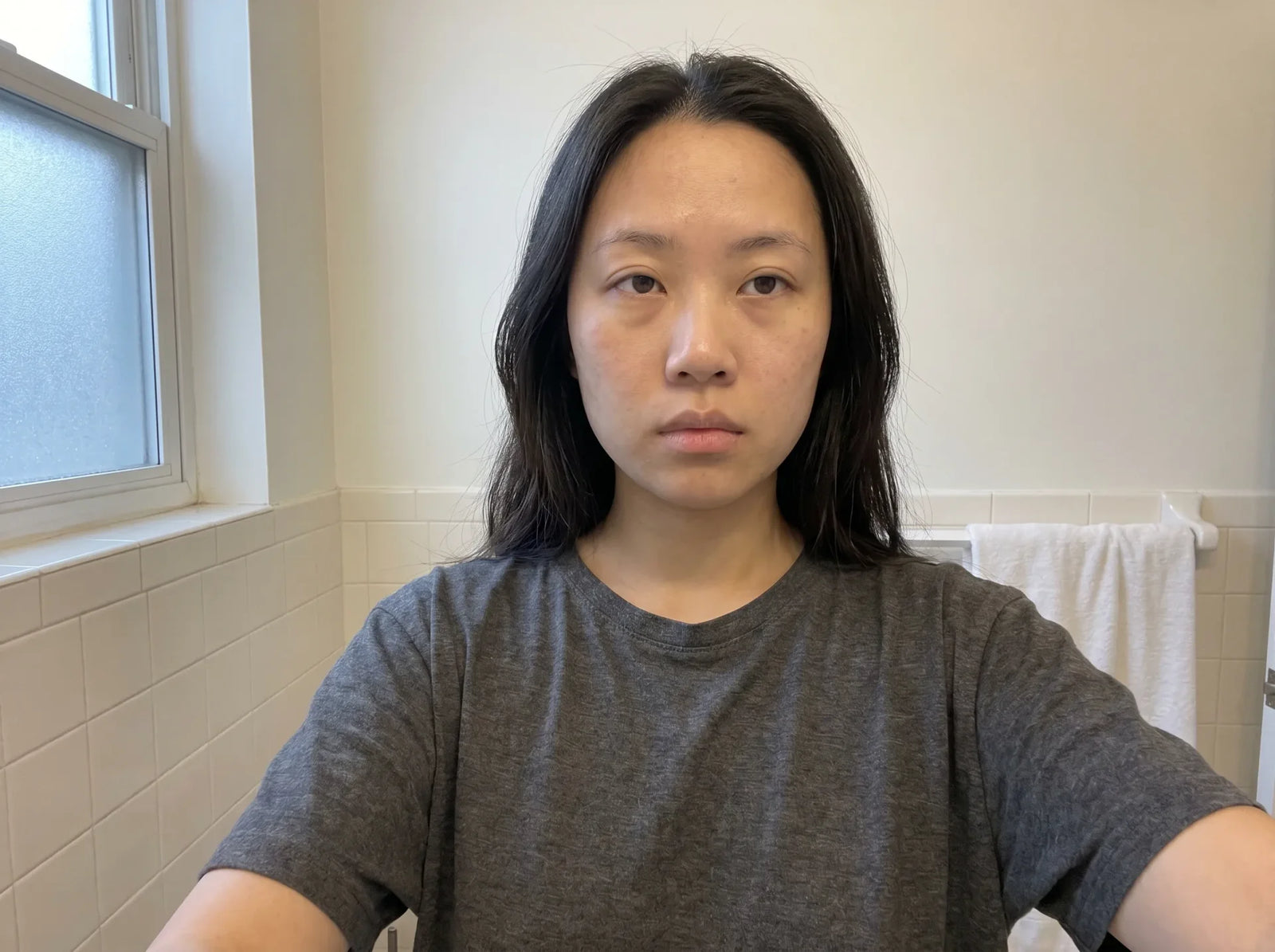 Close-up selfie of a young woman with puffy under-eyes and dull skin, no makeup, wet hair, gray t-shirt, bathroom setting