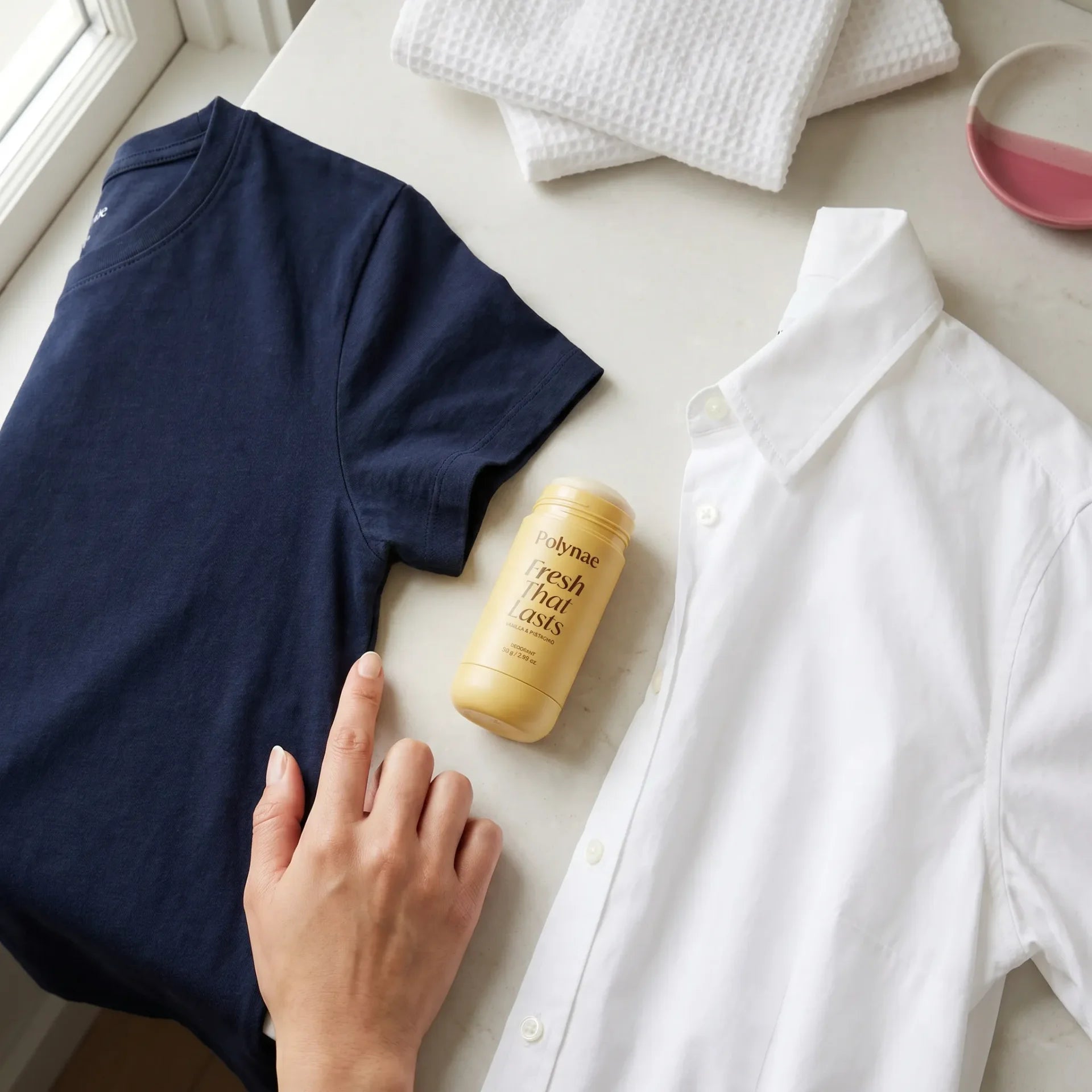 Hand resting on a folded navy t-shirt beside Polynae Fresh That Lasts deodorant in vanilla and pistachio, next to a white button-down shirt on a marble surface