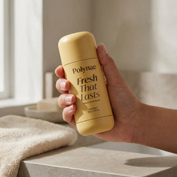 Hand holding a yellow Polynae Vanilla & Pistachio deodorant stick in a minimal bathroom setting with a folded towel nearby.