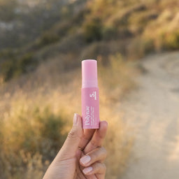 Hand holding a single Polynae pink balm stick up against a warm golden-hour outdoor hillside landscape
