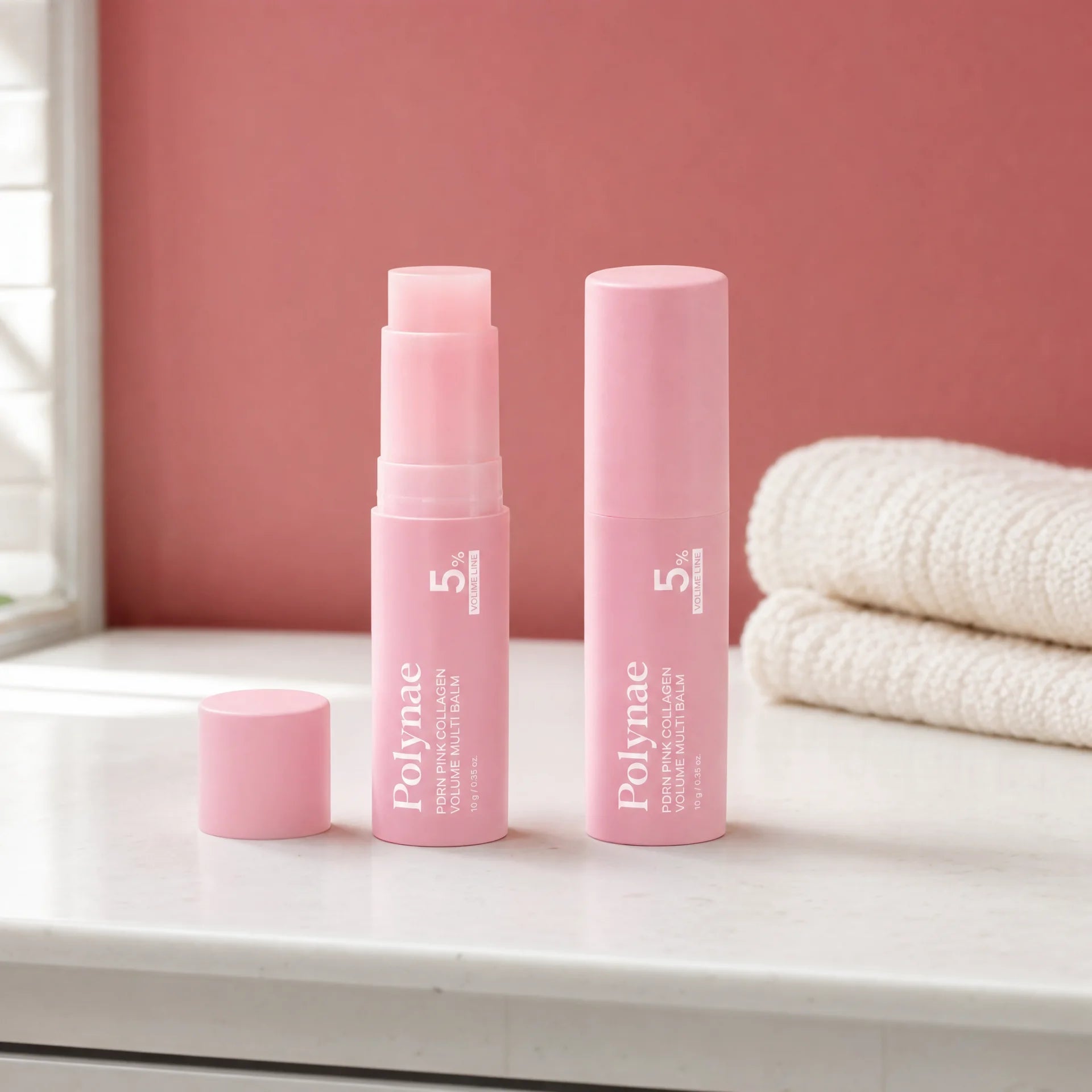 Two Polynae PDRN Pink Collagen Volume Multi Balm sticks side by side on a white marble surface, one open and one capped, with folded towels in the background