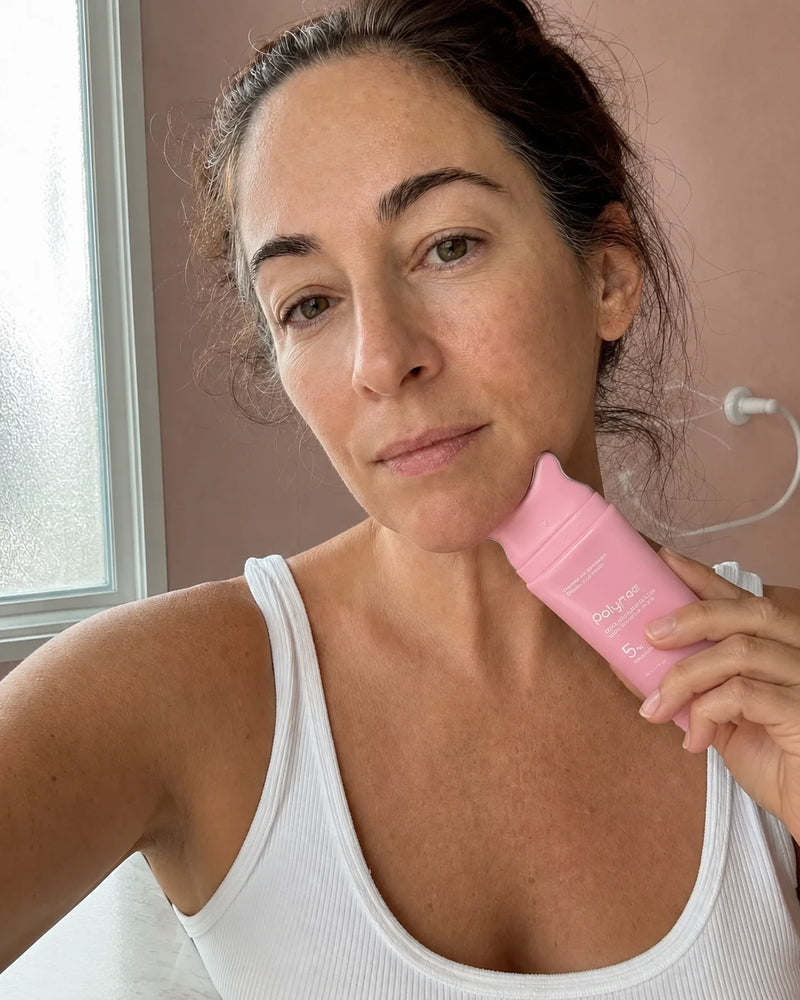 Woman holding a pink Polynae PDRN Collagen Neck Cream tube against her jaw in a bathroom selfie