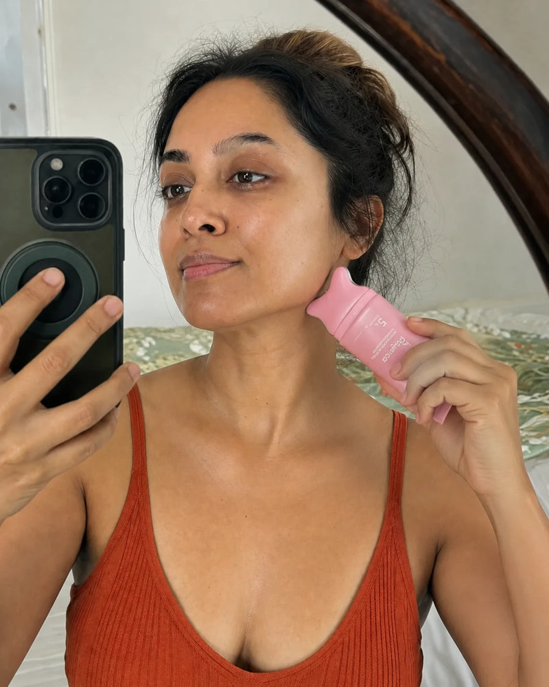 Woman applying a pink Polynae PDRN Collagen Gua Sha Neck Cream roller to her jawline while taking a mirror selfie