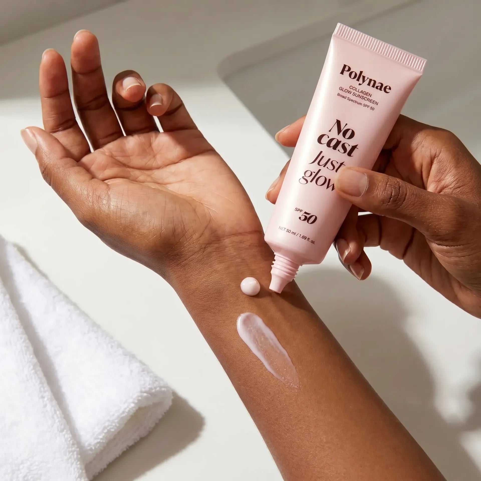A small amount of Polynae Collagen Glow Sunscreen SPF 50 being squeezed from a pink tube onto a dark-skinned forearm, showing a nearly clear, sheer swatch with 