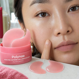 Asian woman with Polynae eye patches applied under both eyes, touching her cheek, with the open product jar on the surface beside her