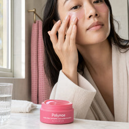 Woman in a cream robe applying a pink Polynae PDRN Caffeine Collagen Eye Patch under her eye at a bathroom vanity, open jar in foreground