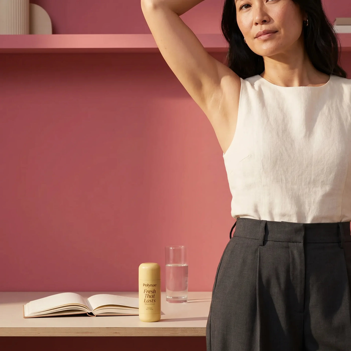 Woman in white linen top raises arm confidently beside Polynae Fresh That Lasts deodorant and an open book on a desk shelf
