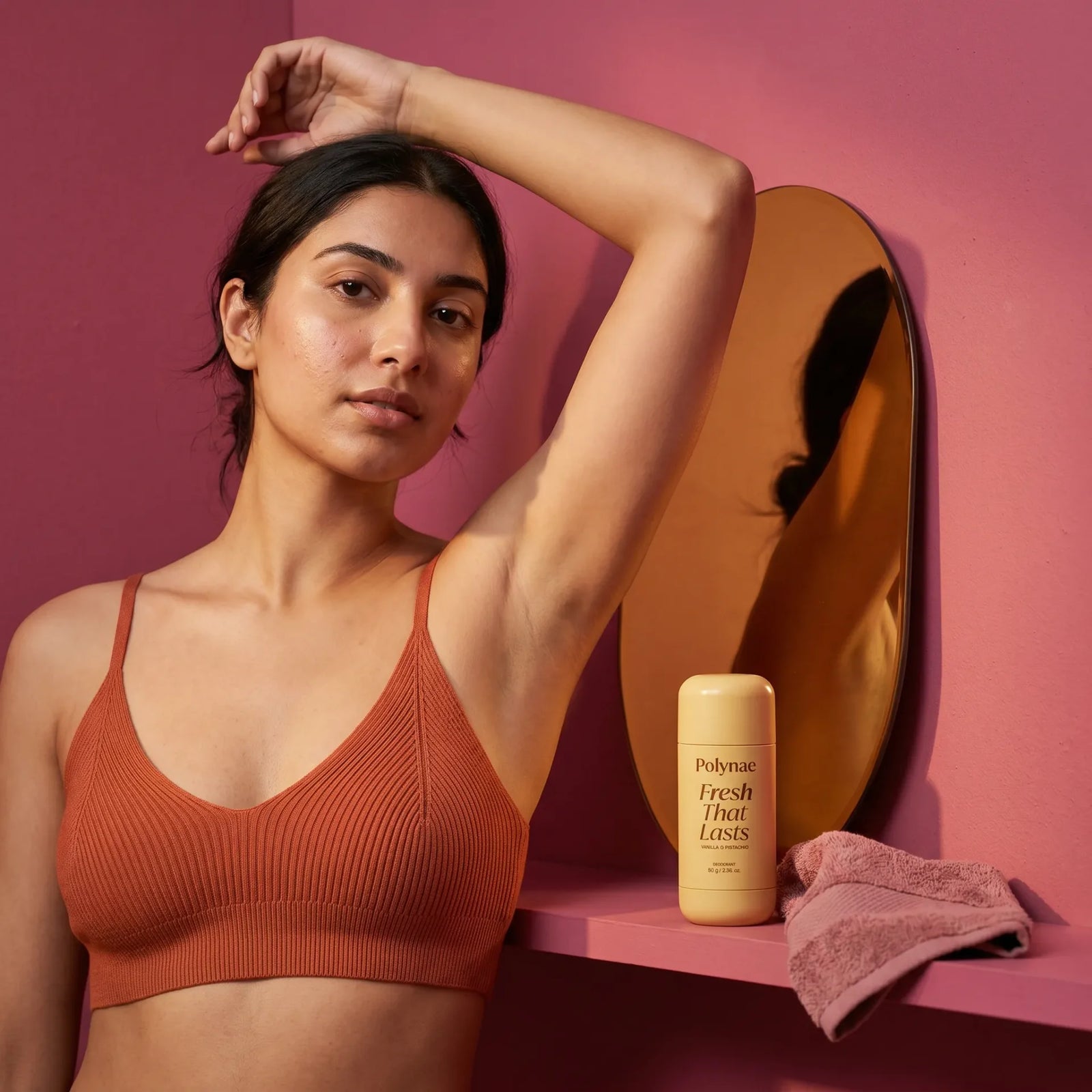 Woman in rust sports bra raises arm next to Polynae Fresh That Lasts deodorant on a pink shelf in front of a round mirror