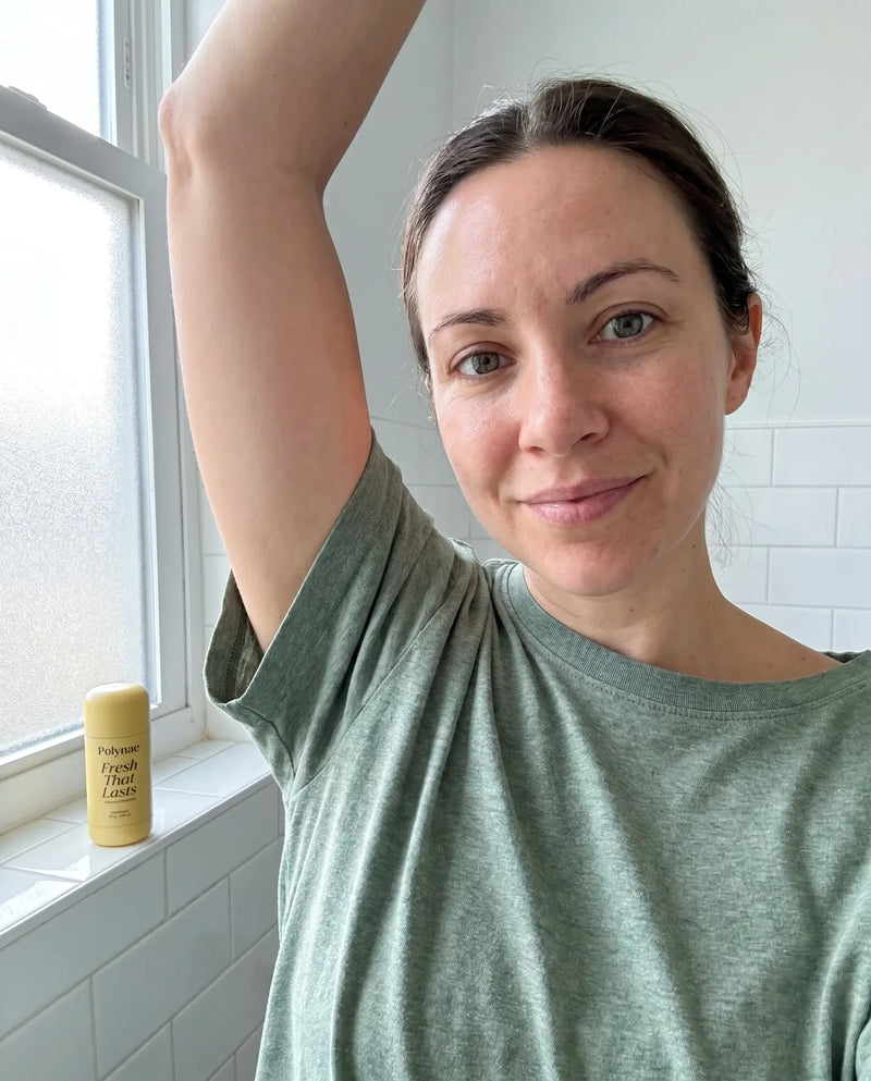 Woman raising her arm in a bathroom to show her underarm, with a yellow Polynae Fresh That Lasts deodorant stick on the windowsill beside her.