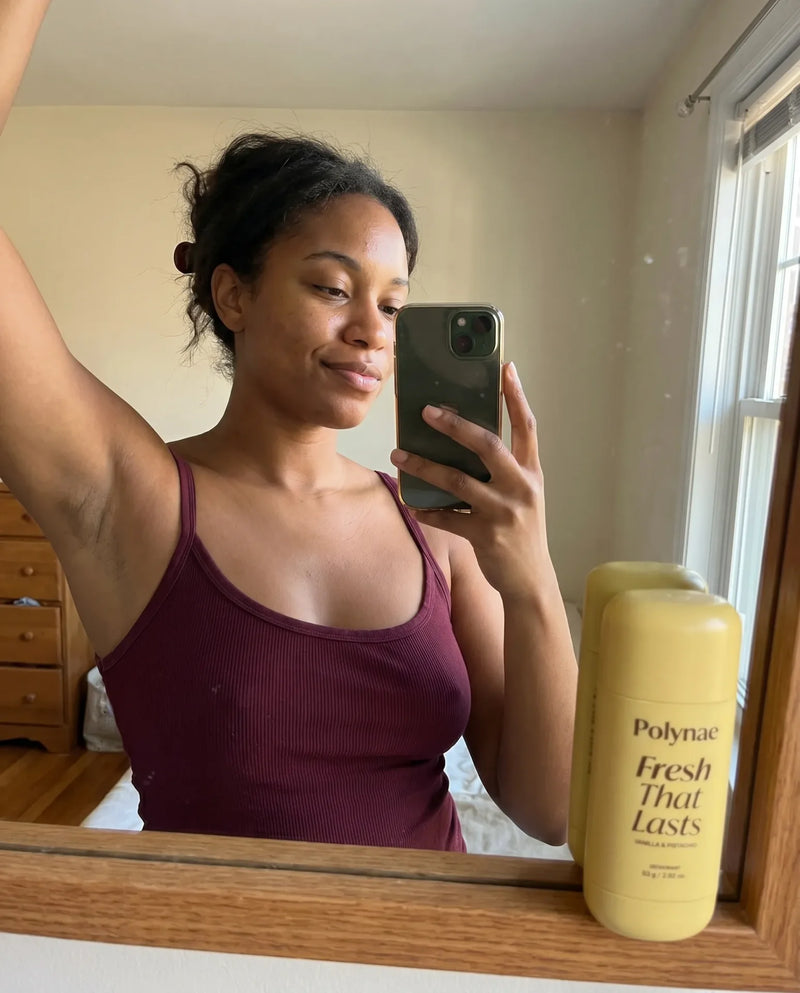 Woman in burgundy tank top takes mirror selfie with arm raised, Polynae Fresh That Lasts deodorant displayed prominently in foreground