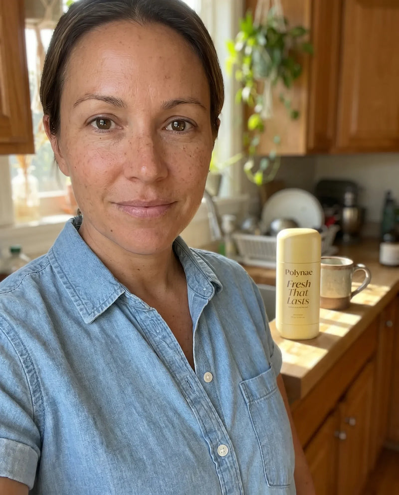 Woman in denim shirt takes selfie in a bright kitchen with Polynae Fresh That Lasts deodorant on the counter beside her