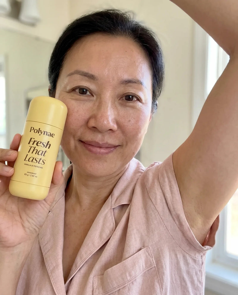 Woman holds Polynae Fresh That Lasts deodorant up toward camera while raising her opposite arm in a bathroom mirror selfie