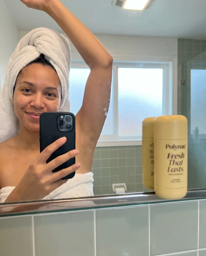 Woman wrapped in a towel after showering takes bathroom mirror selfie with arm raised, two Polynae Fresh That Lasts deodorant sticks on the shelf