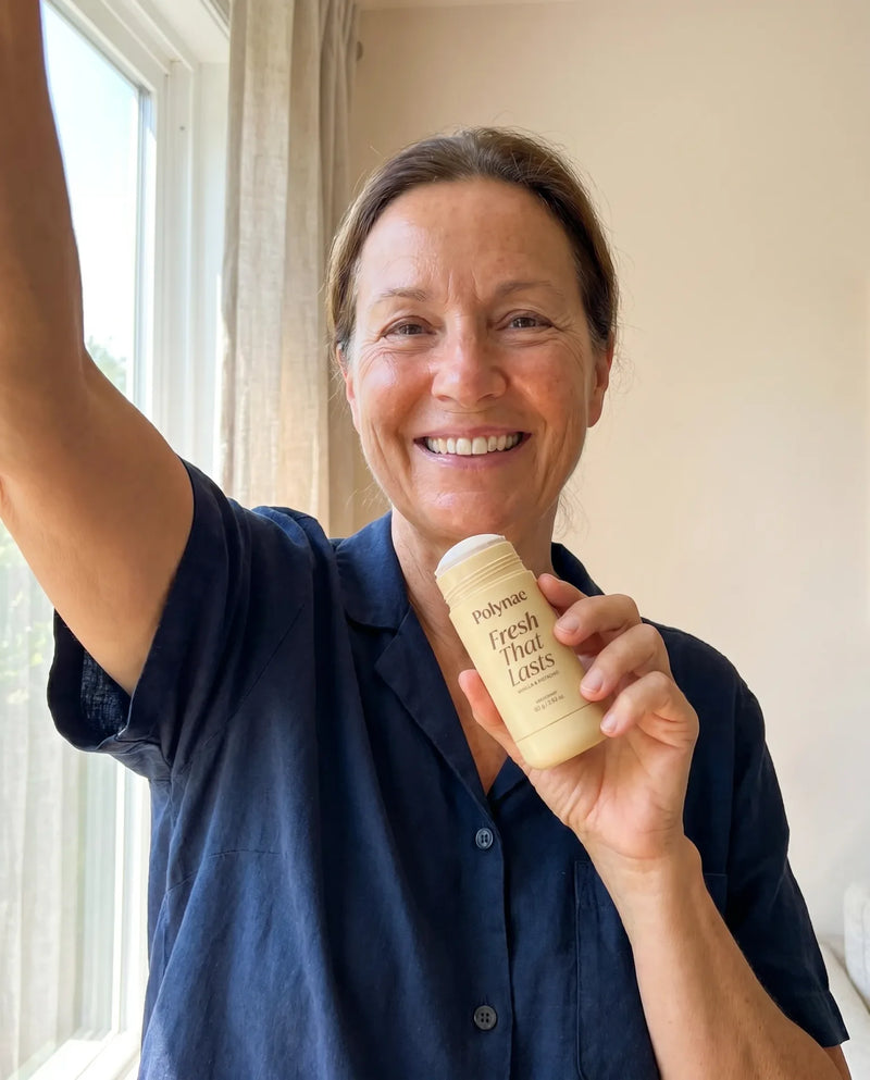 Smiling woman raising one arm and holding a Polynae Fresh That Lasts deodorant stick, vanilla and pistachio scent, near a sunny window