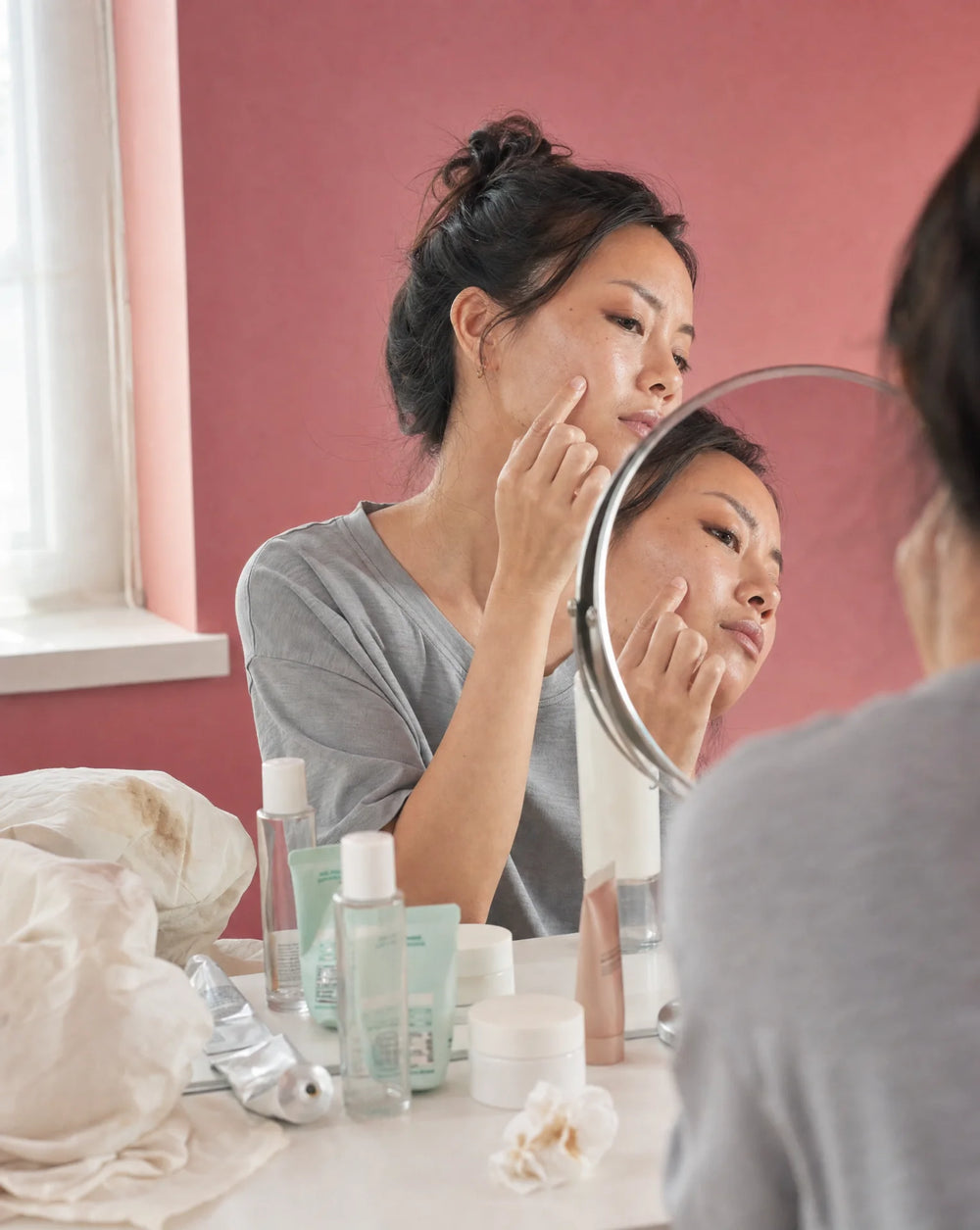 Woman in gray t-shirt pressing her fingertip into her cheek, examining skin concerns in a small round mirror, multiple skincare products scattered on the table