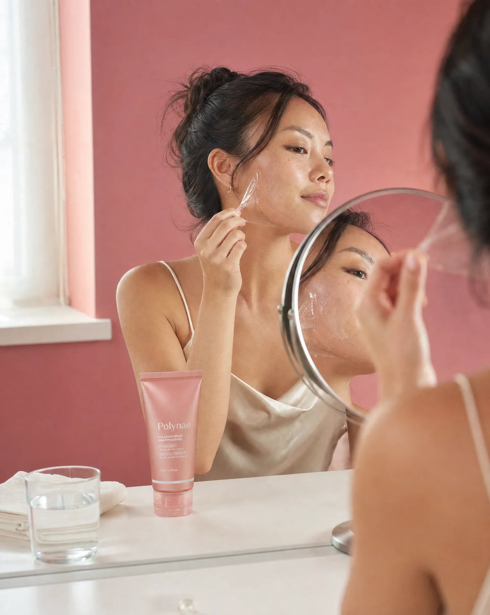 Woman in silk camisole peeling a translucent film mask off her cheek while looking in a round mirror, Polynae tube on the vanity