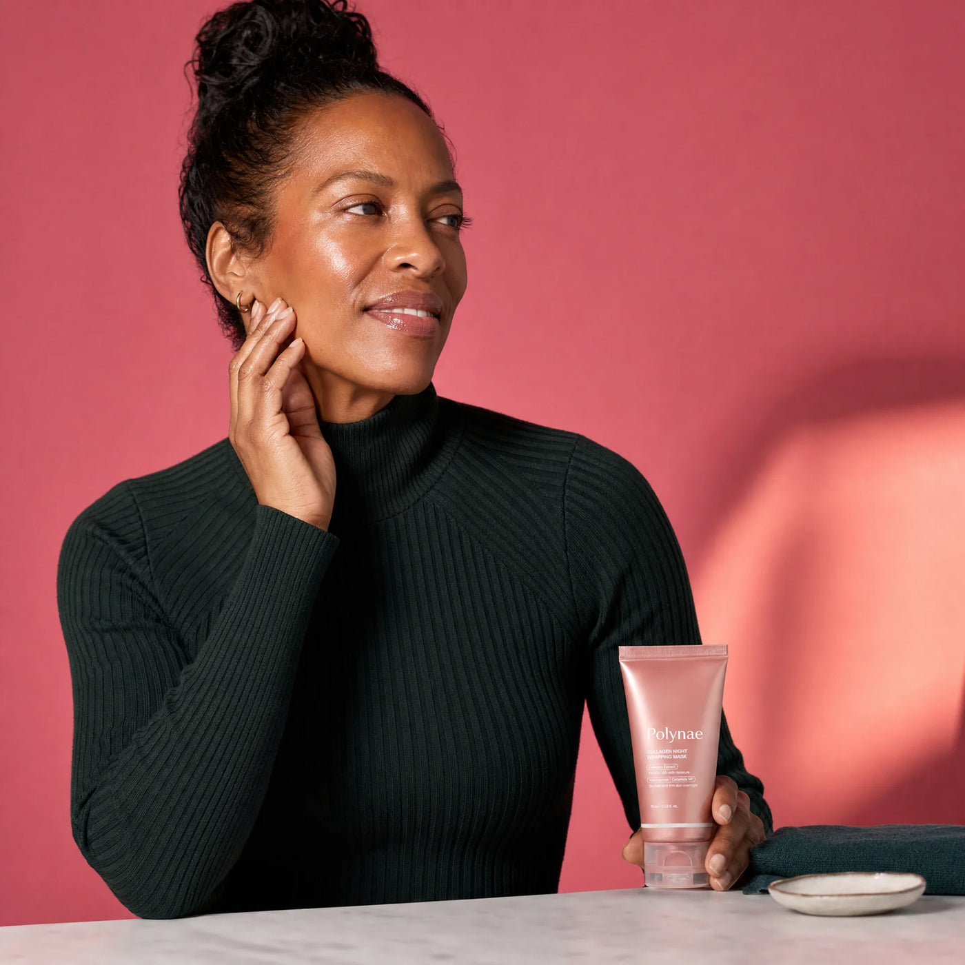 Woman in dark turtleneck holding a Polynae Collagen Night Wrapping Mask tube beside her glowing cheek, hand resting lightly on her jaw