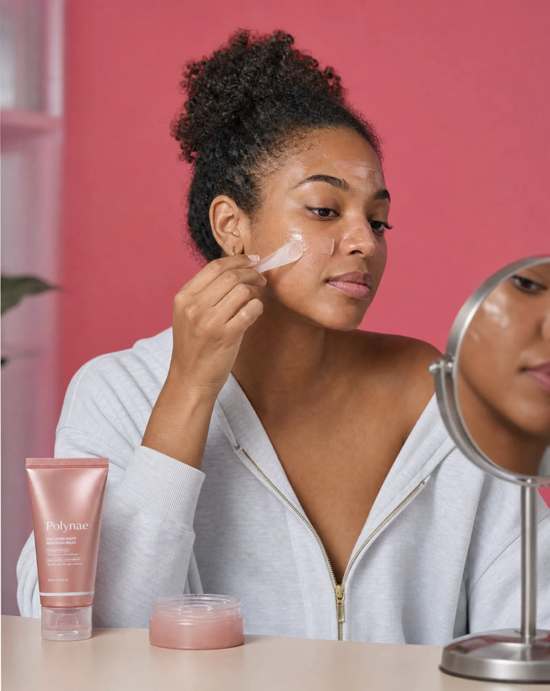 Woman in a white zip hoodie spreading a translucent pink mask from a small jar onto her cheek with a flat spatula, Polynae tube beside a round mirror
