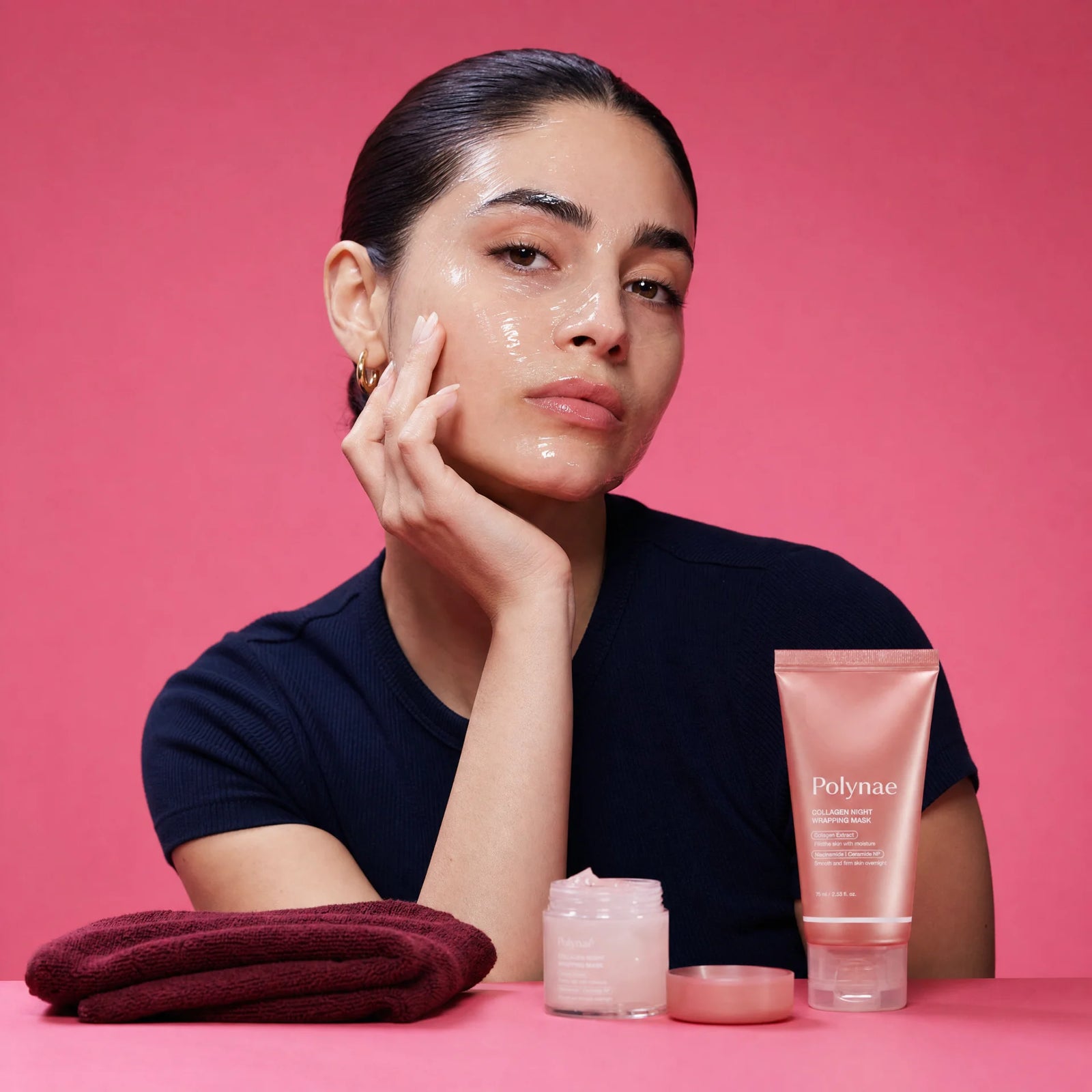 Woman in navy t-shirt with a glossy translucent mask coating her face, resting her chin on her hand, Polynae tube and open jar on the table