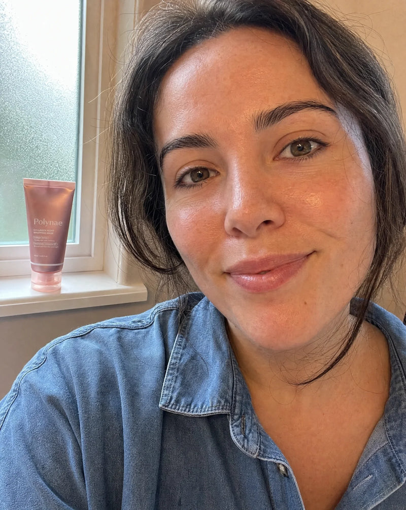 Selfie of a woman with dark hair and hazel eyes in a denim shirt, glowing bare skin, Polynae tube on the windowsill behind her