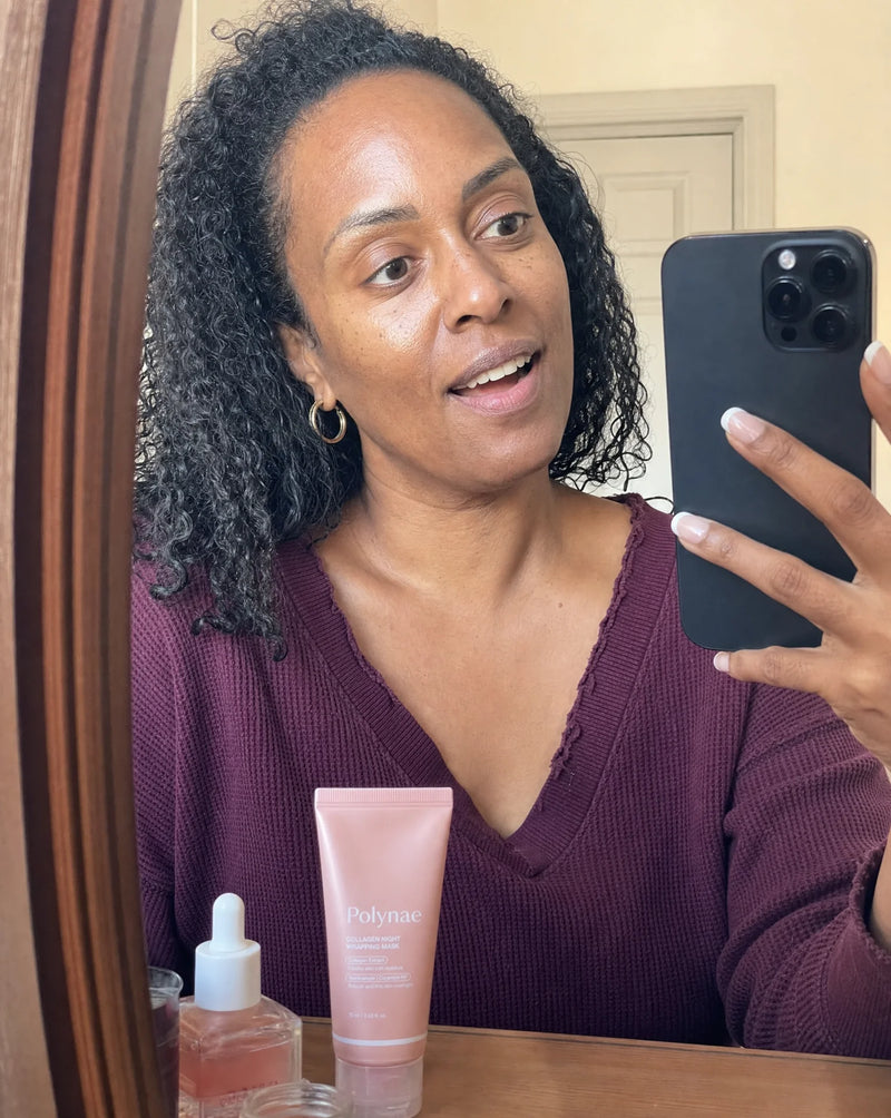 Mirror selfie of a woman in a burgundy v-neck sweater holding a phone, Polynae tube and a dropper serum on the counter in front of her