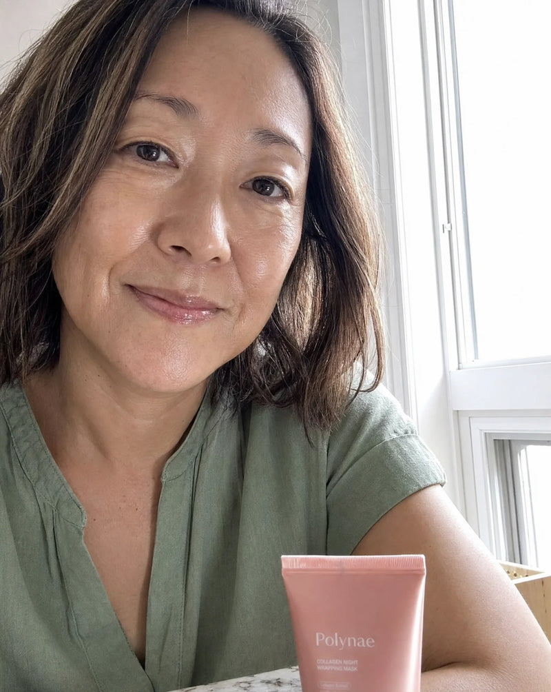 Selfie of a woman with shoulder-length dark hair in a green top holding the Polynae tube near a sunlit window, smooth complexion