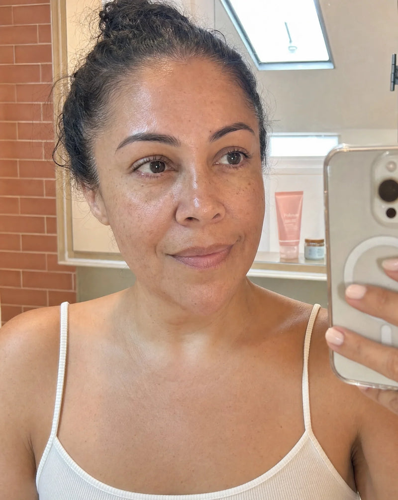 Mirror selfie of a woman in a white tank top with the Polynae tube on a bathroom shelf behind her, dewy skin under warm light