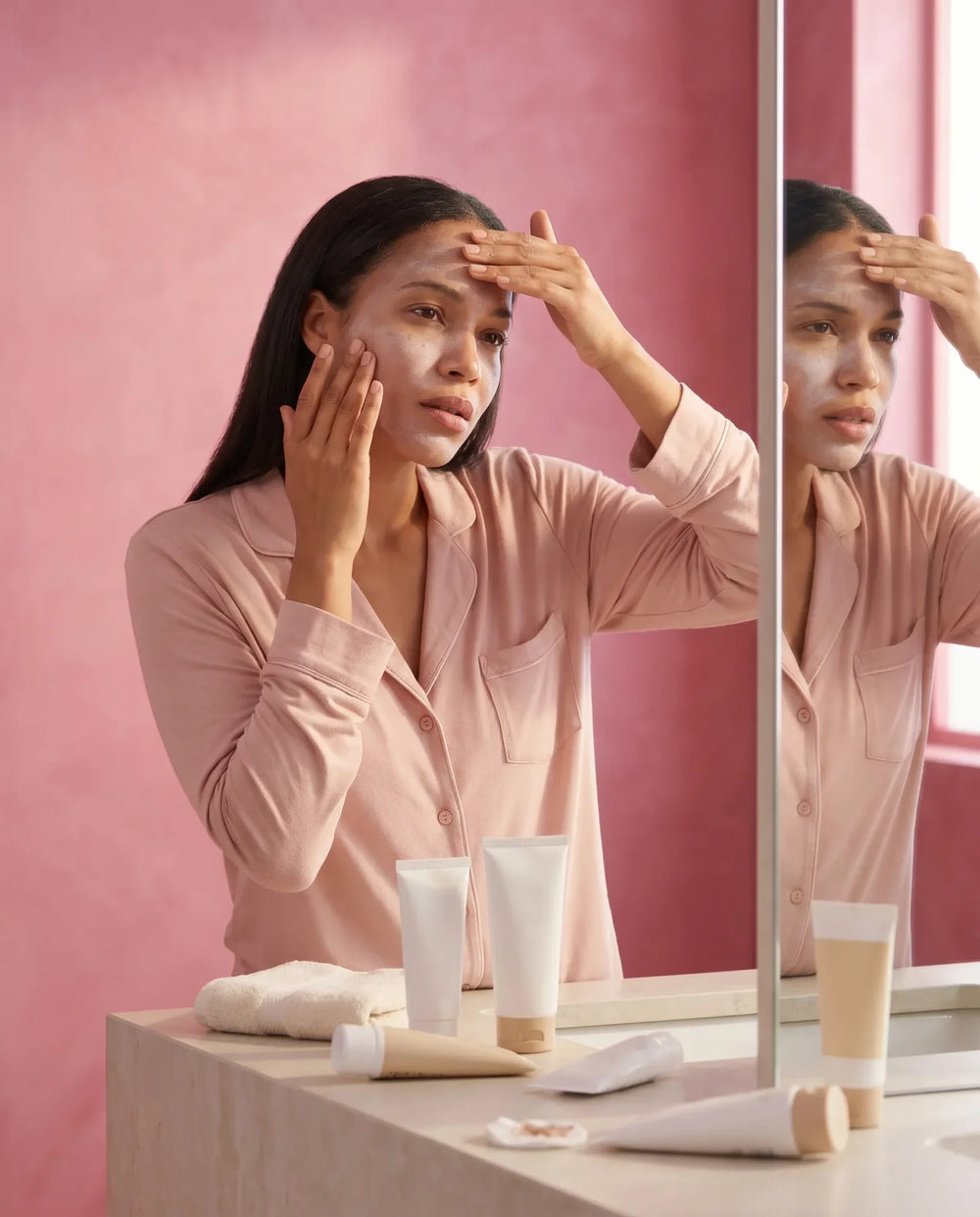 Woman spreading thick white cream across her forehead and cheek, visible white residue on skin, multiple unlabeled skincare tubes on counter