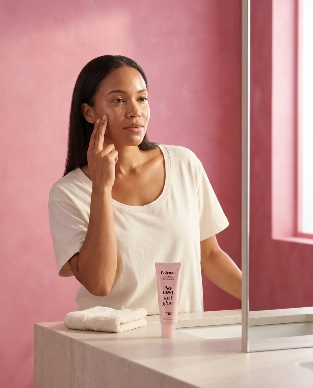 Woman applying Polynae No Cast Just Glow SPF 50 sunscreen to her cheek at a bathroom vanity, product tube beside a folded towel
