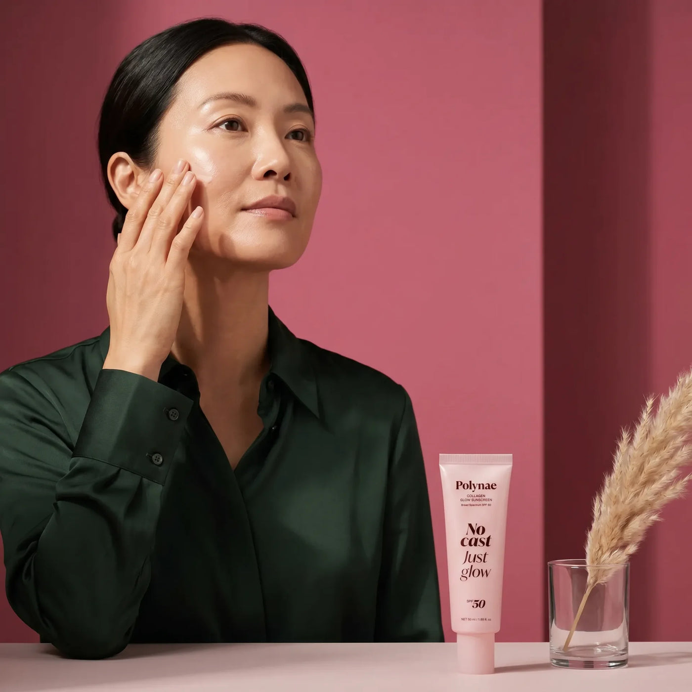 Woman in dark green shirt touching her cheek beside a Polynae No Cast Just Glow SPF 50 tube and a glass vase with dried pampas grass