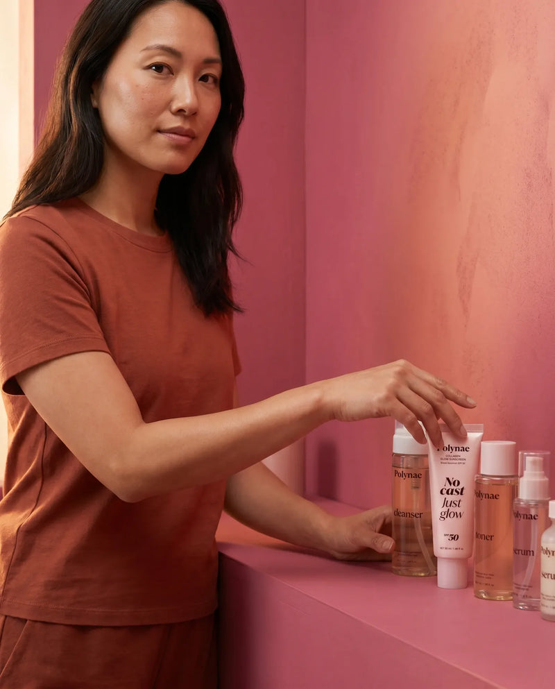 Woman arranging Polynae skincare products including cleanser, toner, and serum on a pink shelf
