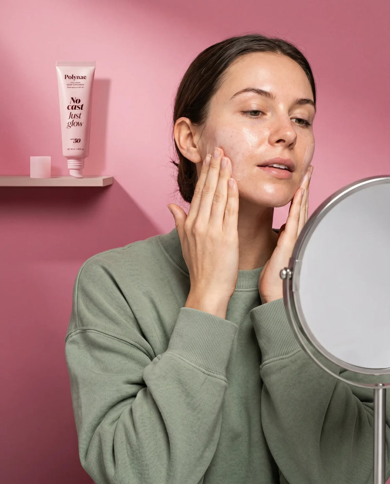 Woman applying Polynae Collagen Glow Sunscreen SPF 50 to her cheek while looking into a round mirror