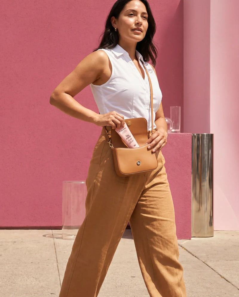 Woman pulling a tube of Polynae No Cast Just Glow SPF 50 out of a tan crossbody bag outdoors