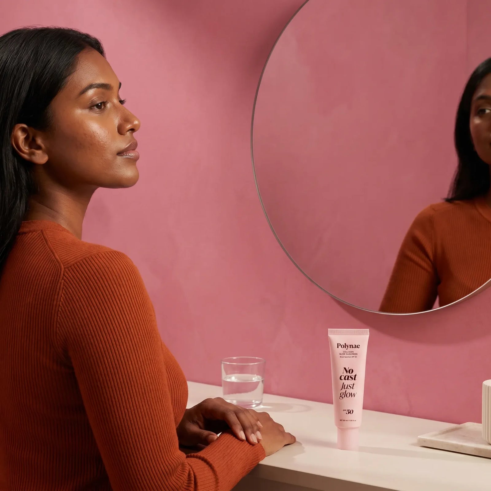 Woman with dark skin sitting at a vanity mirror, no white cast visible after wearing Polynae Collagen Glow Sunscreen SPF 50