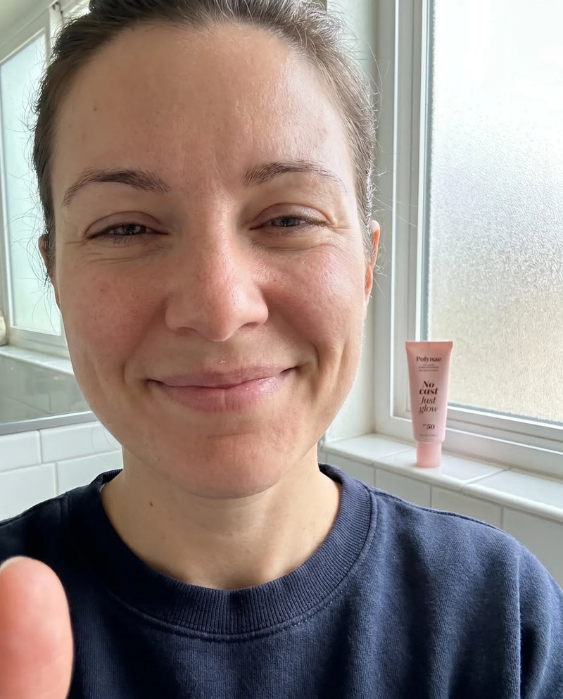 Candid bathroom selfie of a smiling woman with light skin wearing a navy sweatshirt, Polynae SPF 50 tube on the windowsill behind her