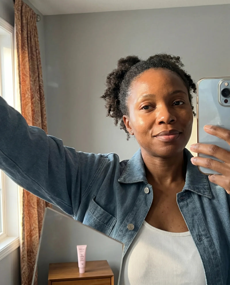 Mirror selfie of a woman with natural hair wearing a denim jacket, Polynae sunscreen tube on a side table, skin appears healthy and even