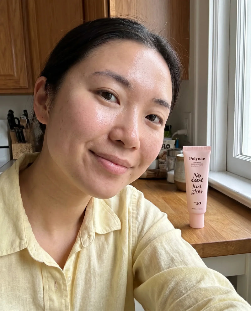 Woman with dark hair in a yellow shirt smiling beside a Polynae No Cast Just Glow SPF 50 tube on a kitchen counter