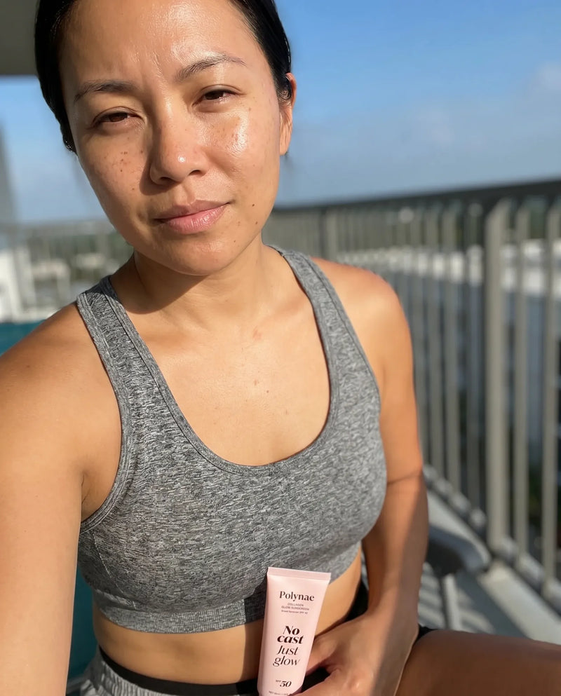 Woman in a gray sports bra on a sunny outdoor balcony holding Polynae SPF 50 sunscreen, slight sweat visible on face