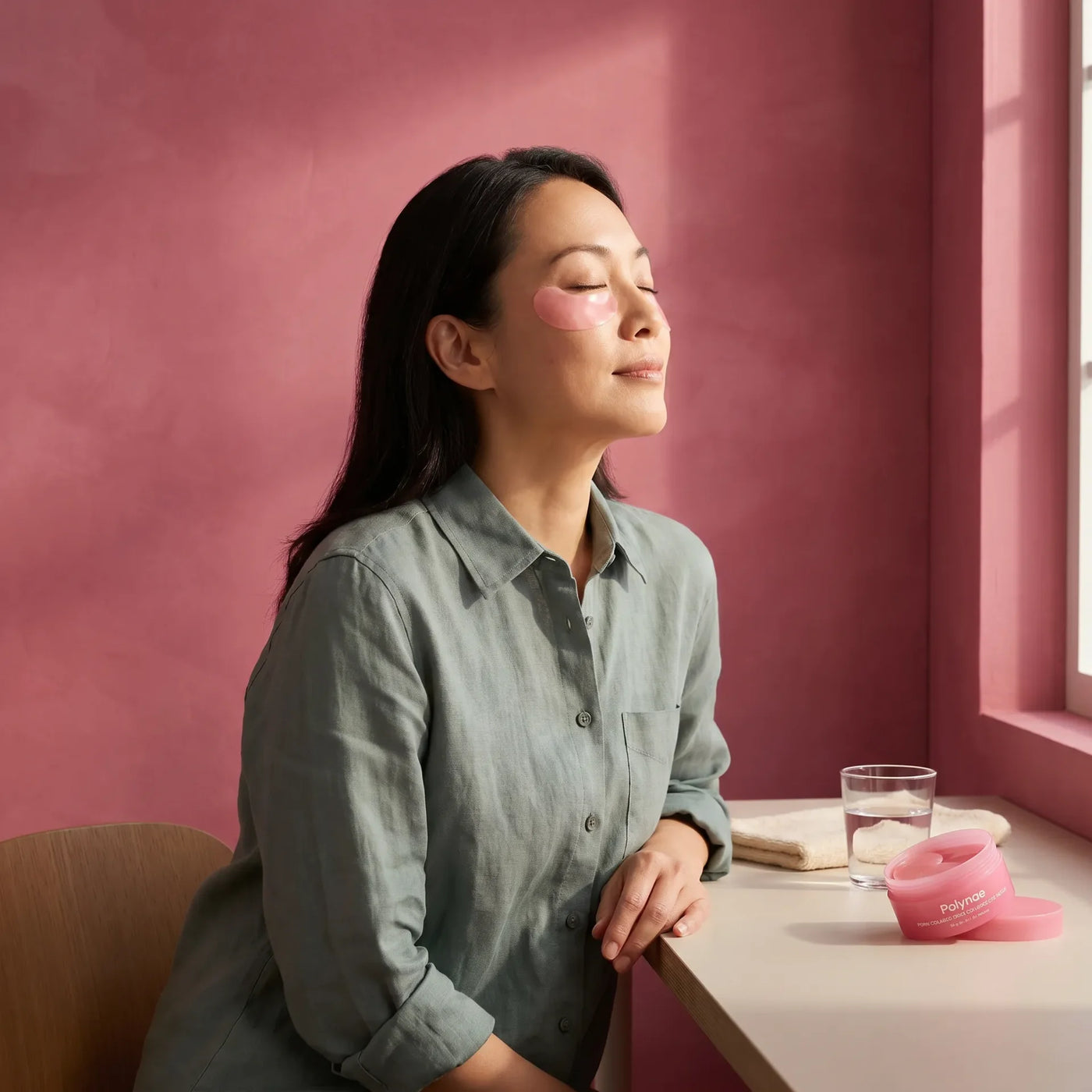 Woman with eyes closed and pink under-eye gel patches applied, seated near a sunlit window beside an open Polynae skincare jar and glass of water