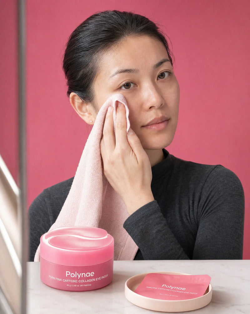 Woman patting her under-eye area with a soft pink towel, seated at a vanity with the Polynae PDRN Pink Caffeine Collagen Eye Patch jar and single patch on a dis