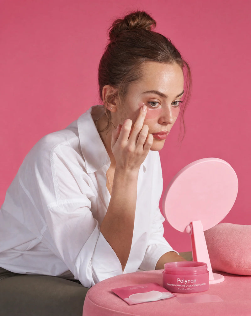 Woman in a white shirt applying a Polynae eye patch under one eye using a finger, small mirror and jar on a pink table