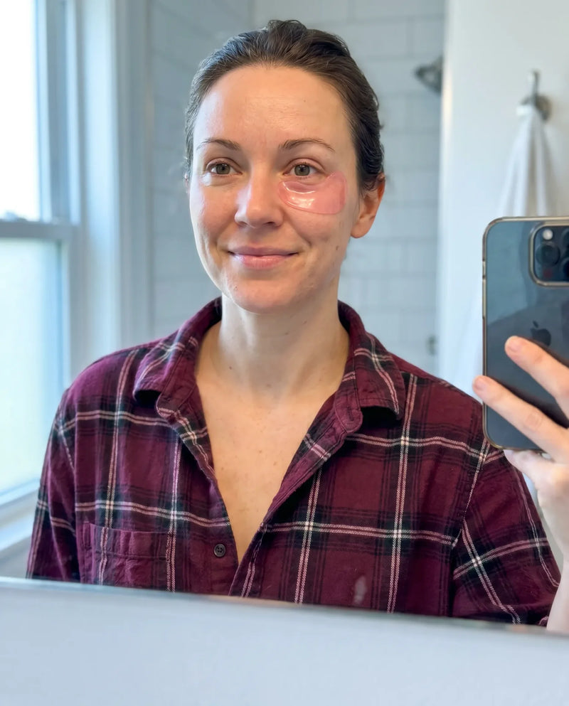 Woman taking a bathroom mirror selfie wearing pink under-eye gel patches on both cheeks and a dark plaid flannel shirt