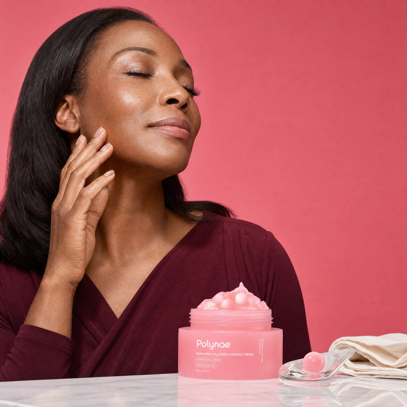 Woman with eyes closed touching her jawline beside an open jar of Polynae Porn Pink Collagen Capsule Cream on a marble surface