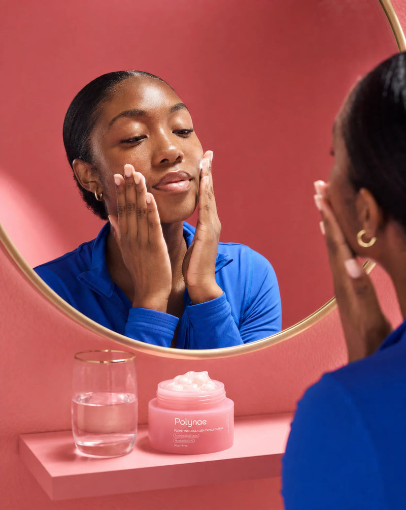 Woman in blue top applying Polynae cream to both cheeks while looking in a round gold-framed mirror