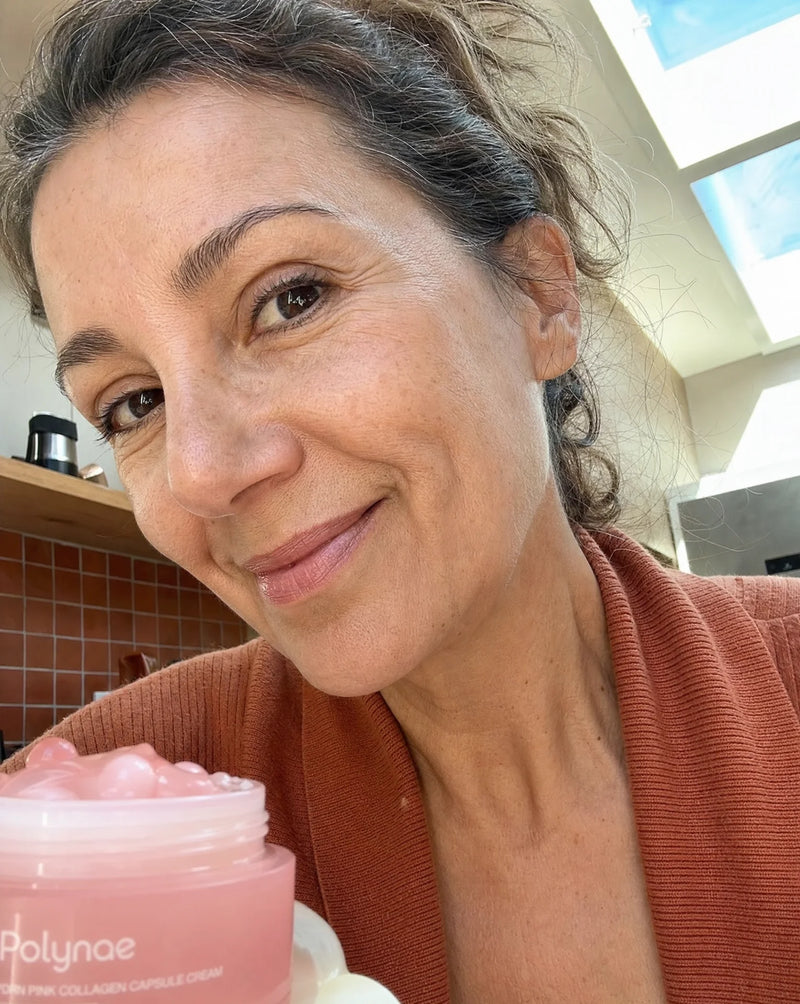 Older woman smiling with the Polynae cream jar in a naturally lit kitchen, skin visibly smooth