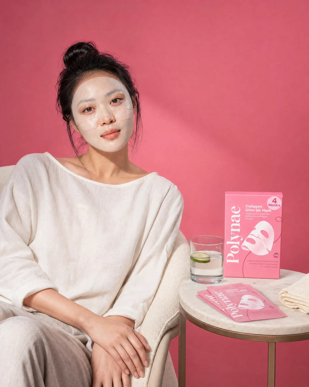 Woman wearing Polynae Collagen Glow Up sheet mask, seated calmly beside a small table with the pink product box and a glass of water