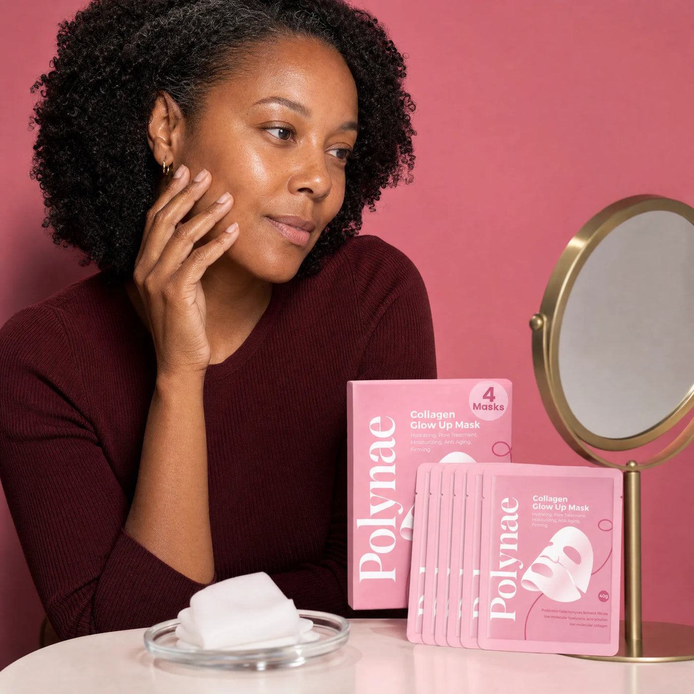 Woman in burgundy top touching her cheek beside Polynae Collagen Glow Up Mask box and individual packets stacked on a table with a round mirror