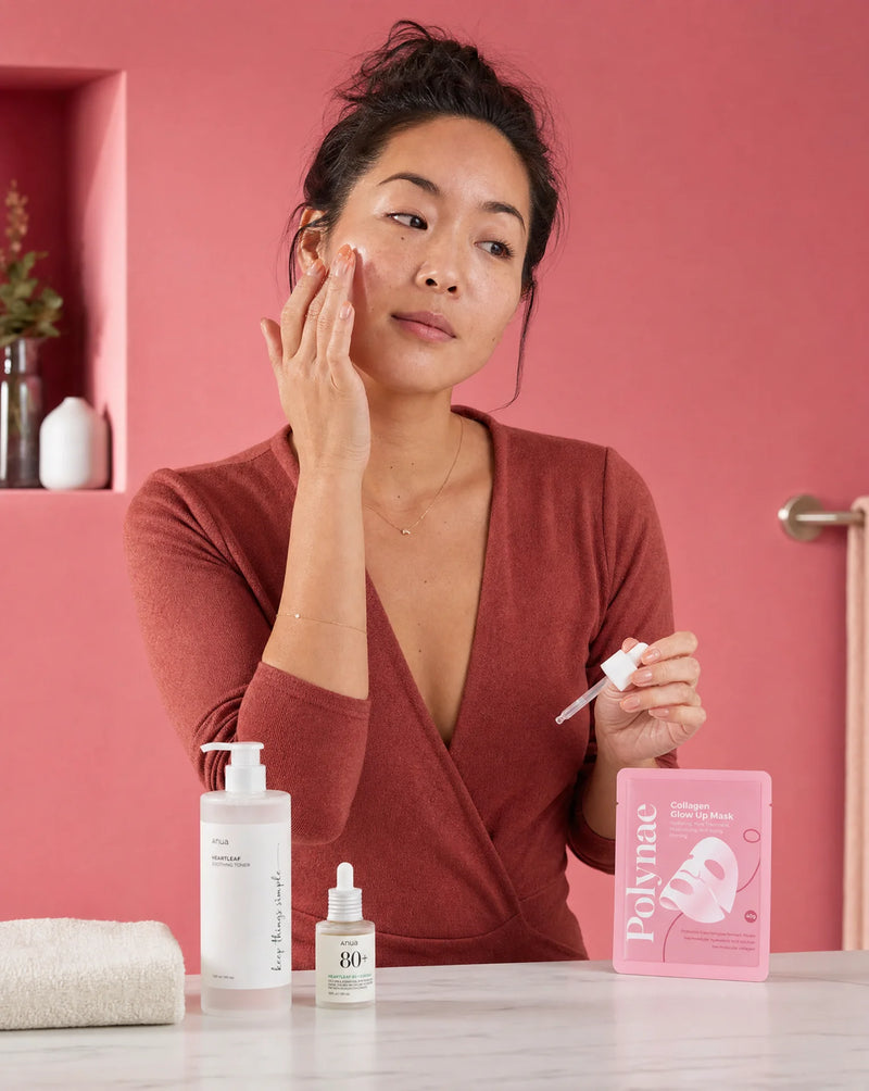 Woman in a rust-colored wrap top applying serum to her face beside Polynae mask packets, an Anua toner, and a dropper bottle on a marble surface