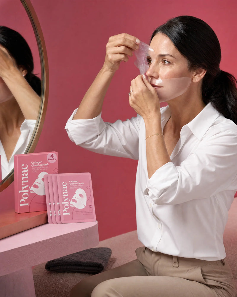 Woman in a white shirt peeling a translucent Polynae sheet mask away from her face while looking in a round mirror, mask packets stacked nearby